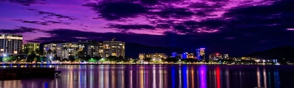 The Cairns city waterfront at dusk, high-rise lights reflecting across the water under a purple sky